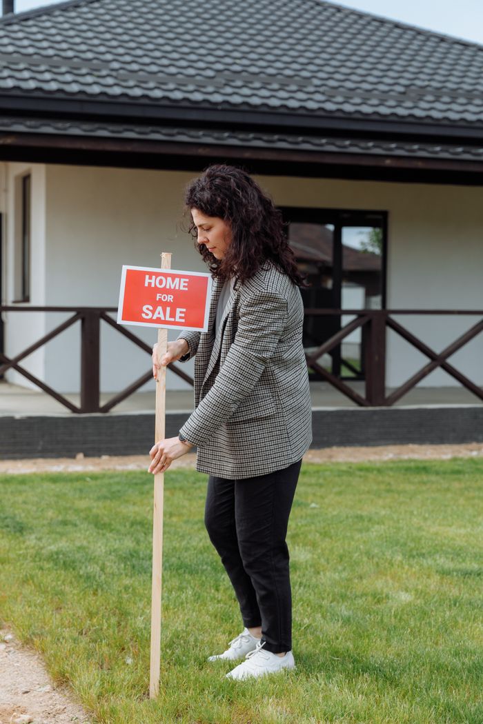 Woman placing a home for sale sign in front of a house, showcasing real estate career.